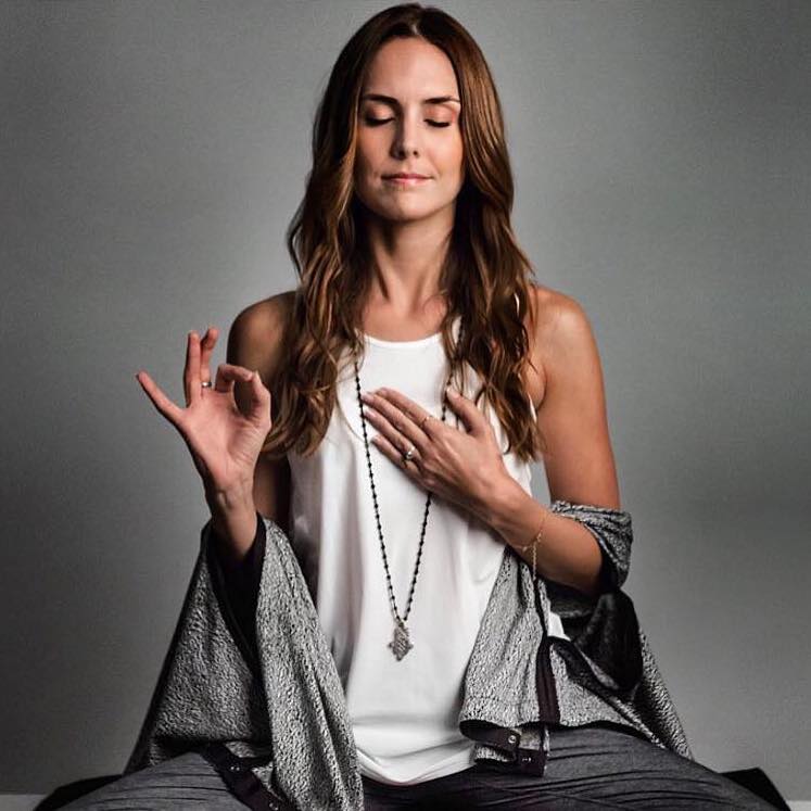 Lauren Eckstrom sits with her right hand in a gyan mudra, her left hand on her heart, and her eyes closed.